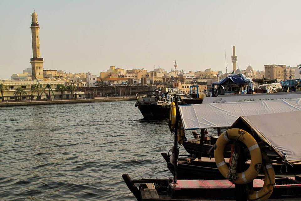 Traditional wooden abra boats moored on Dubai Creek, with the old Bur Dubai waterfront and a mosque minaret across the water in warm, hazy light